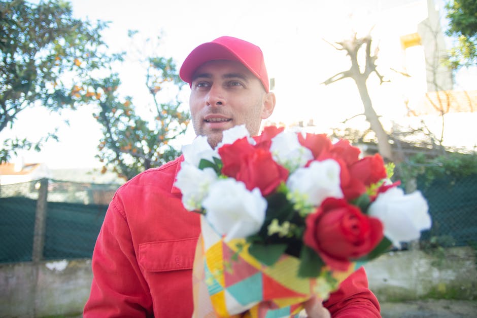 A male flower delivery person wearing a red cap and matching red jacket holds a vibrant floral bouquet towards the camera outdoors. The bouquet features a mix of red and white roses, with green foliag