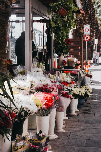 A street view outside a florist shop on a cloudy day, featuring an array of fresh flower bouquets arranged in white vases and buckets along the sidewalk. The floral compositions include white roses, p