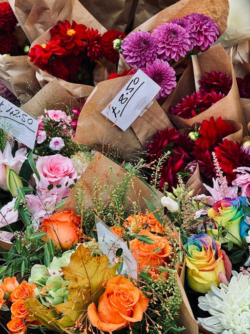 A vibrant display of fresh flowers arranged in paper wrapping at a floral market stall. The bouquet features bright orange roses with lush green foliage, complemented by soft pink roses and delicate w