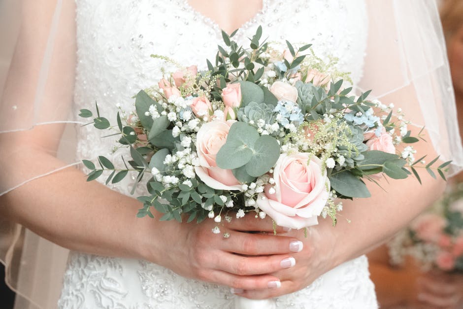 A bride in a white lace dress holding a bridal bouquet with soft pink roses, white baby's breath, eucalyptus leaves, and greenery. The bouquet is arranged in a rounded style with the flowers and folia
