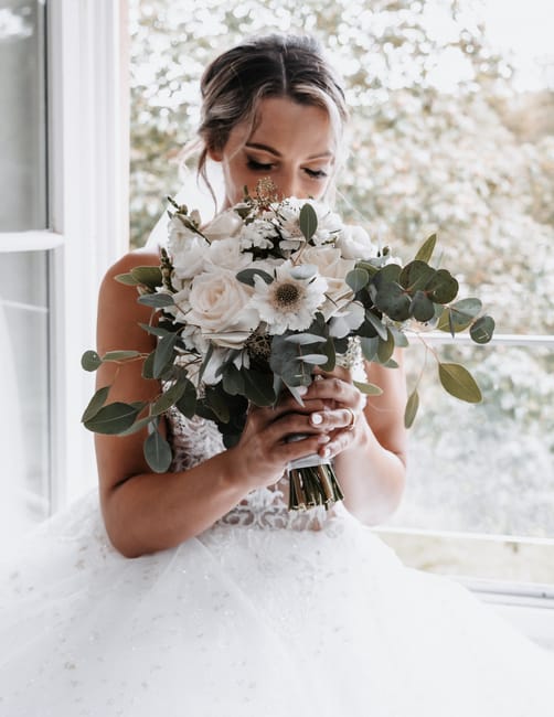 A bride holds a large, elegant bouquet featuring cream and white flowers, including roses, anemones with dark centers, and delicate filler blooms, accented with silvery-green eucalyptus leaves. The bo