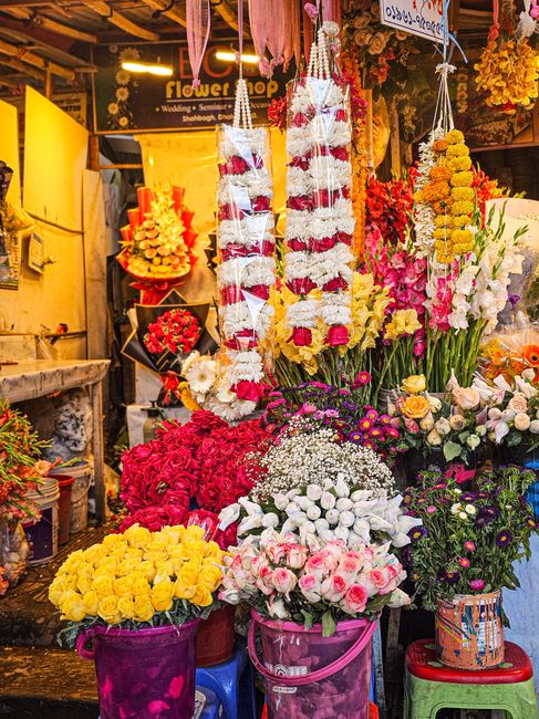 A vibrant flower stall filled with fresh floral arrangements and bouquets in a variety of colours. Prominent in the display are large garlands made of white and red flowers, hanging vertically. Surrou