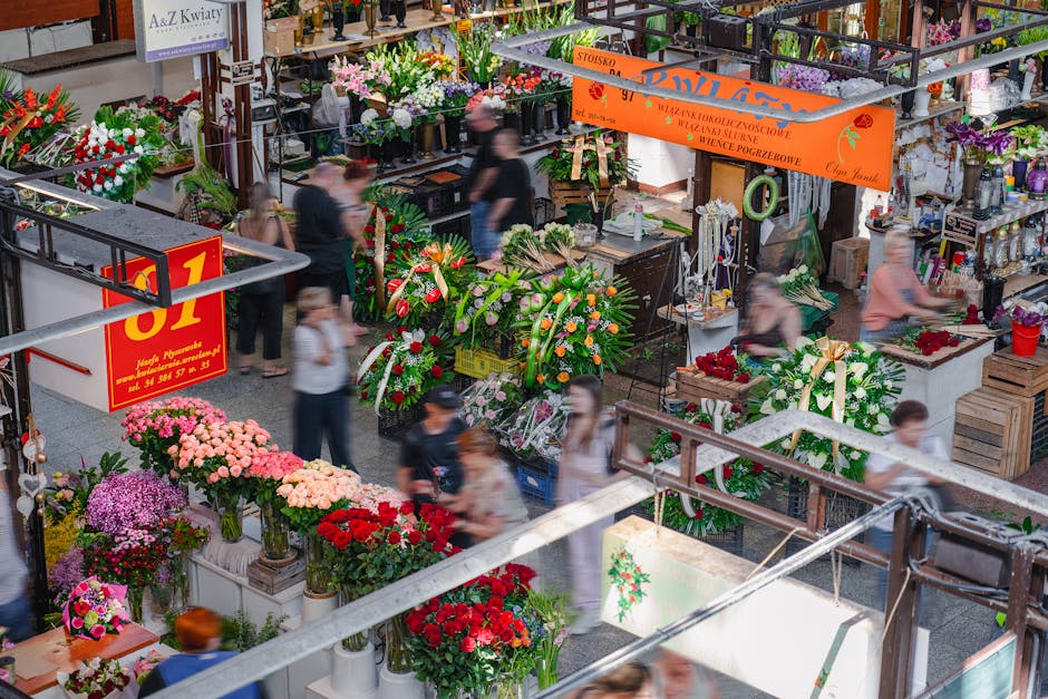 A bustling indoor flower market displaying a vibrant assortment of fresh floral arrangements and bouquets. In the foreground, numerous potted chrysanthemums in shades of pink, purple, and red are arra