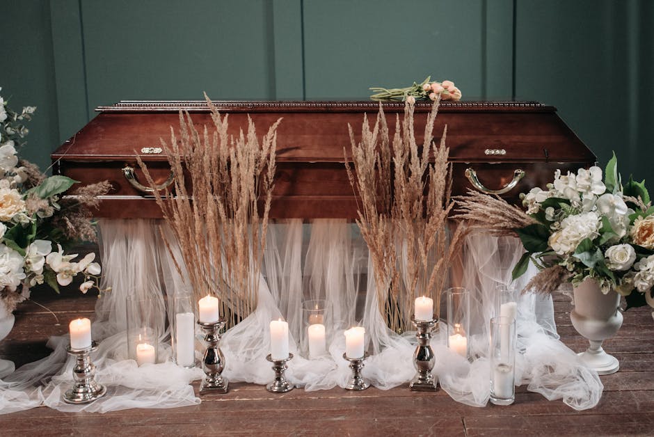 A wooden funeral casket is decorated with fresh floral arrangements on either side, featuring white roses, cream-colored carnations, and greenery, complemented by tall, feathery pampas grass stems pla