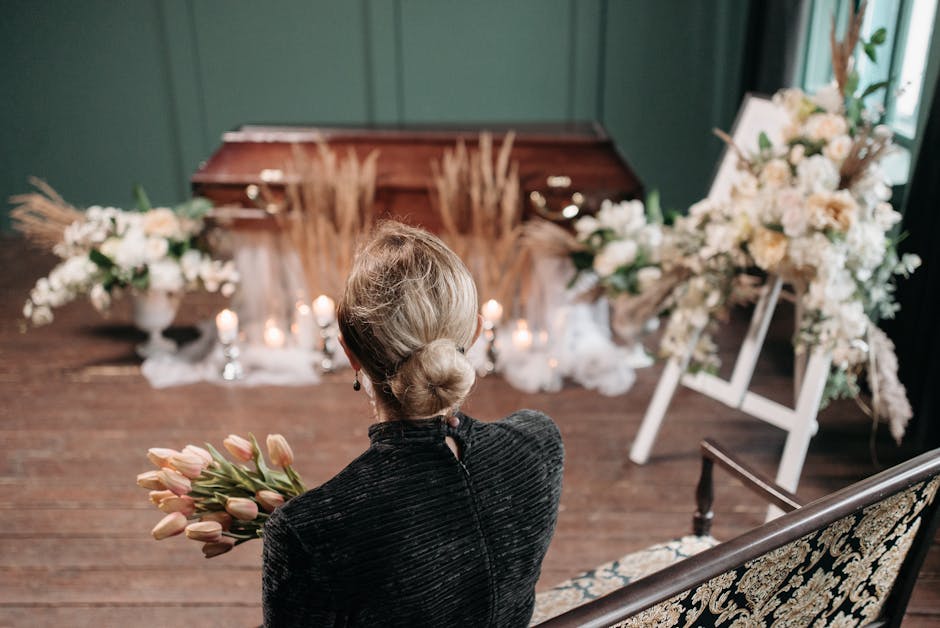 A woman with her hair styled in a low bun, seen from behind, holds a bouquet of pale pink tulips. She is dressed in a dark velvet jacket and is seated on an ornate, vintage-style bench with a floral u