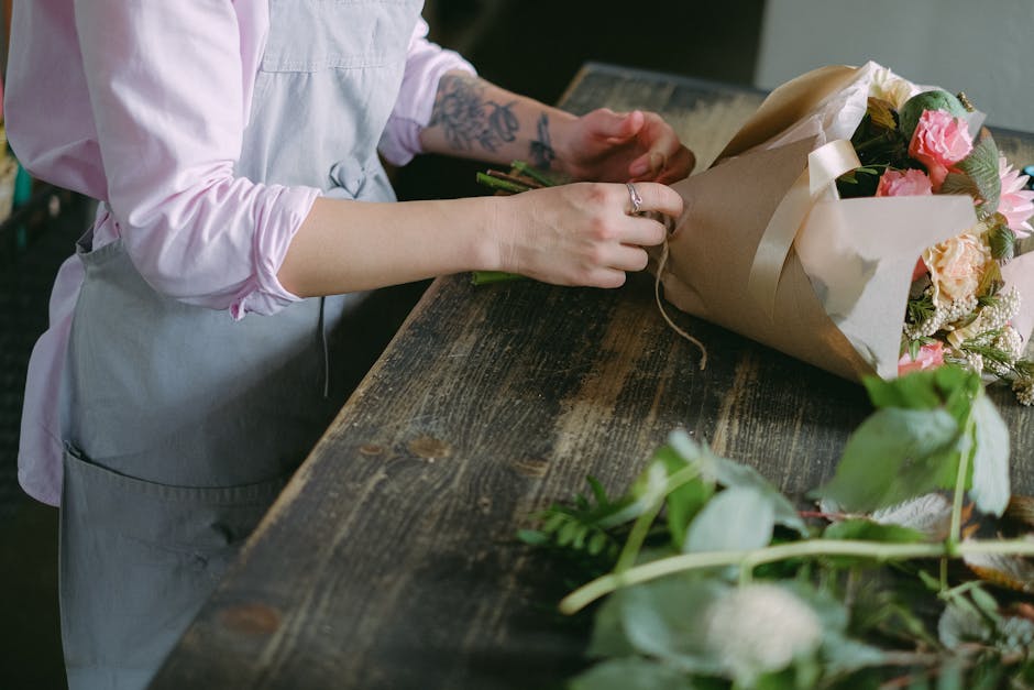 A florist, wearing a light pink shirt and a grey apron, is arranging a bouquet of fresh flowers on a rustic wooden table. The bouquet features pink and peach roses, white and green accents, and is wra