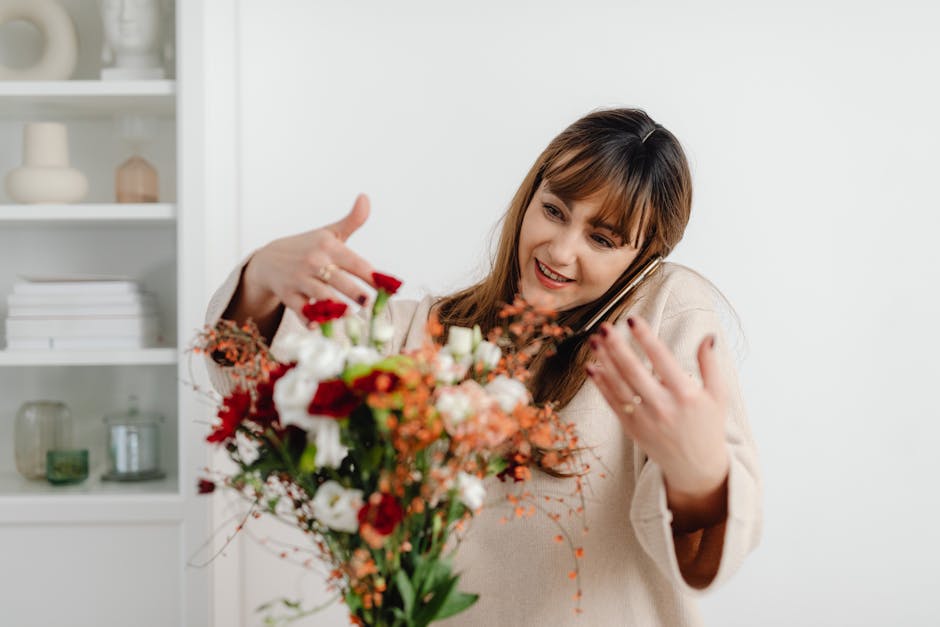 A woman with shoulder-length brown hair, smiling and wearing a beige sweater, is holding a vibrant floral bouquet in her left hand while talking on her smartphone held to her right ear. The bouquet fe